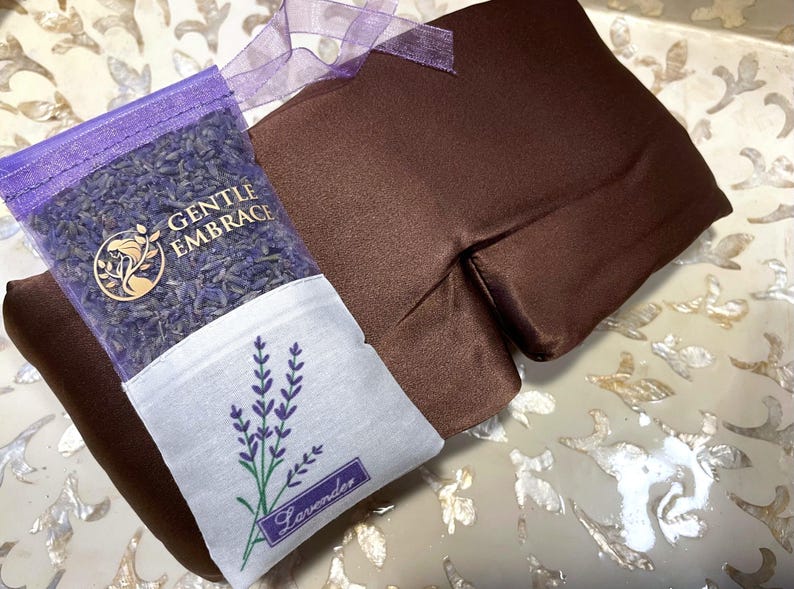 May include: A brown eye pillow and a lavender sachet. The eye pillow is rectangular and made of a smooth fabric. The sachet is filled with lavender buds and has the text "GENTLE EMBRACE" and a lavender sprig design. A purple ribbon is attached.