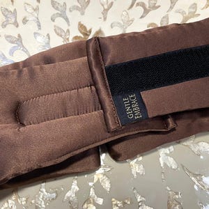 May include: A brown eye pillow with a black Velcro fastening and the text "GENTLE EMBRACE" on a small label. The pillow is made of a soft, satin-like material and is designed to be worn around the eyes.