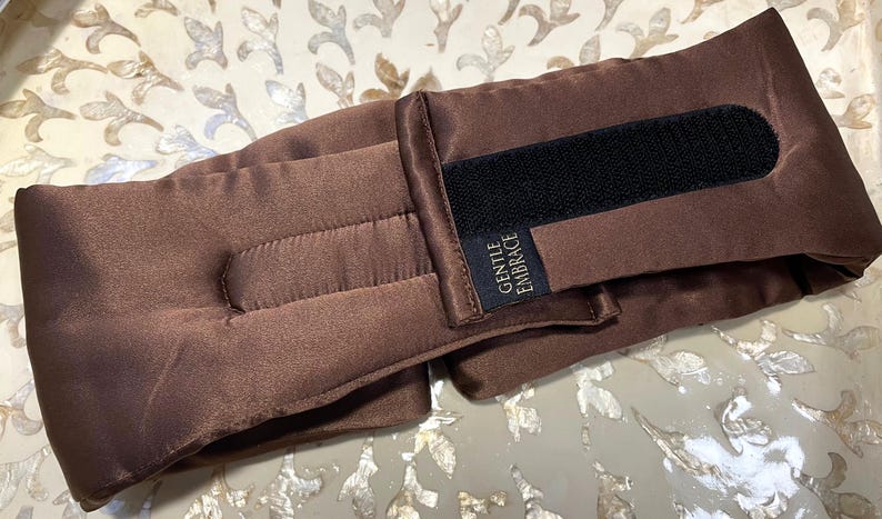May include: A brown eye pillow with a black Velcro fastening and the text "GENTLE EMBRACE" on a small label. The pillow is made of a soft, satin-like material and is designed to be worn around the eyes.