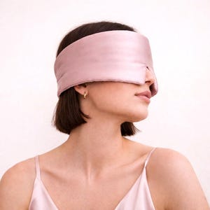 May include: A pale pink sleep mask made from a soft, silky fabric. The mask covers the eyes and is worn around the head. The sleep mask is a solid colour and appears to be made of satin or silk.
