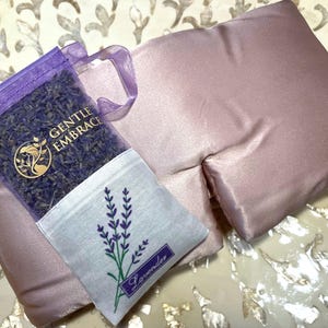 May include: A lavender-scented eye pillow and a sachet of dried lavender. The eye pillow is a soft, dusty rose colour. The sachet is purple with the words "GENTLE EMBRACE" and a lavender sprig design. The sachet also has the word "Lavender".