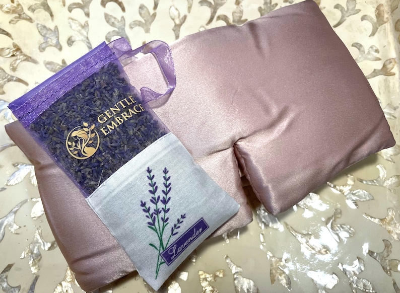 May include: A lavender-scented eye pillow and a sachet of dried lavender. The eye pillow is a soft, dusty rose colour. The sachet is purple with the words "GENTLE EMBRACE" and a lavender sprig design. The sachet also has the word "Lavender".