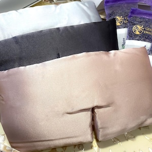 May include: An assortment of eye pillows in white, black, and blush pink, alongside several small bags of dried lavender. The eye pillows are rectangular and made of a smooth, satin-like material. The lavender bags have a purple mesh top.