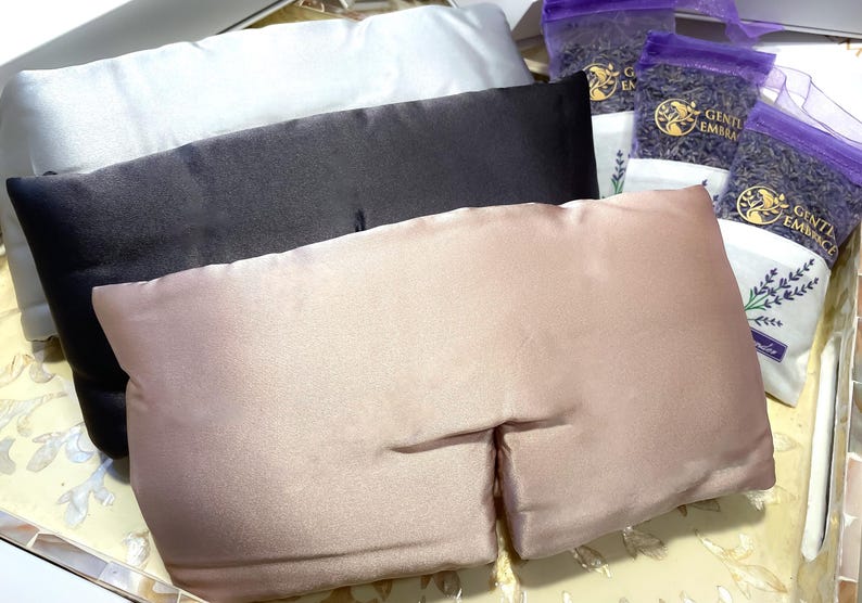 May include: An assortment of eye pillows in white, black, and blush pink, alongside several small bags of dried lavender. The eye pillows are rectangular and made of a smooth, satin-like material. The lavender bags have a purple mesh top.