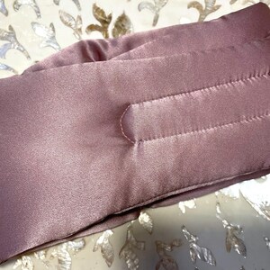 May include: A mauve satin eye mask with a quilted design. The mask features horizontal stitching and a curved nose section. The fabric has a subtle sheen, and the mask appears soft and comfortable, ideal for sleep.