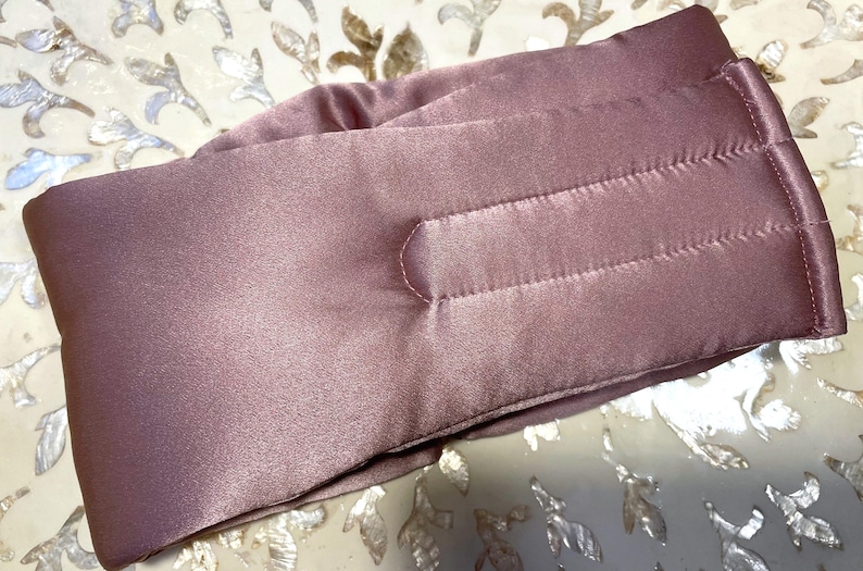 May include: A mauve satin eye mask with a quilted design. The mask features horizontal stitching and a curved nose section. The fabric has a subtle sheen, and the mask appears soft and comfortable, ideal for sleep.