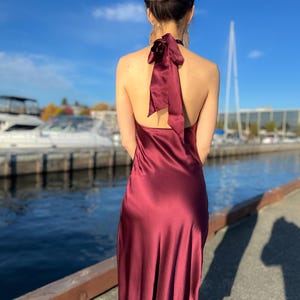 May include: A burgundy satin halter dress with a bow detail at the back. The dress is floor-length and has a flowing silhouette. The background features a blue sky, water, and boats.