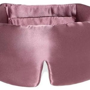 May include: A dusky pink sleep mask crafted from a silky fabric. The mask features a broad band for the head and a shaped eye area. The design is simple and elegant, suggesting a focus on comfort and light blockage for better sleep.