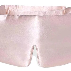 May include: A pale pink satin eye mask. The mask has a rectangular shape with a curved top and a split at the bottom for the nose. The material looks soft and smooth, suggesting comfort. The eye mask is designed to block out light.