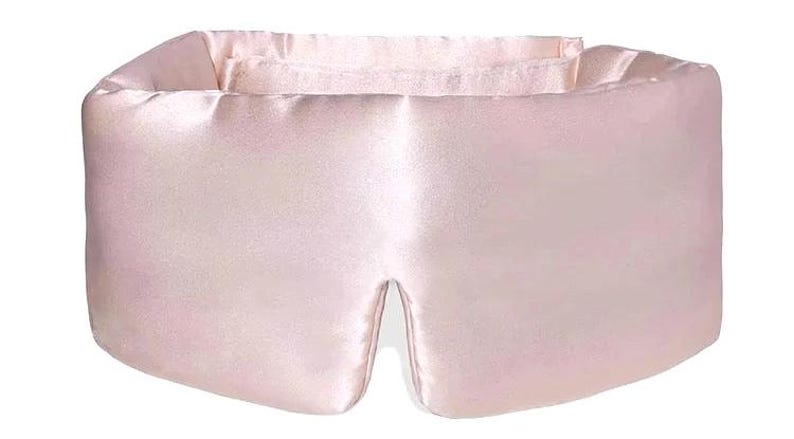 May include: A pale pink satin eye mask. The mask has a rectangular shape with a curved top and a split at the bottom for the nose. The material looks soft and smooth, suggesting comfort. The eye mask is designed to block out light.