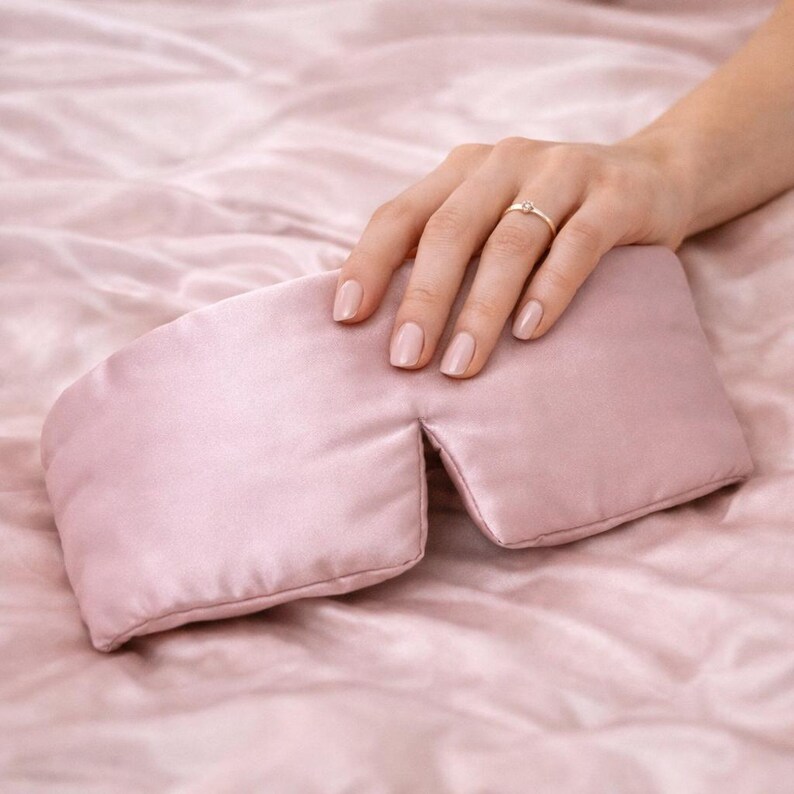 May include: A pale pink satin eye mask. The mask is rectangular with a curved shape. The mask is on a pink satin surface. A hand with a ring rests on the mask. The eye mask is designed to block out light.