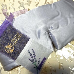 May include: A light grey satin eye pillow and a small sachet filled with dried lavender. The sachet has a lavender sprig illustration and the word "Lavender". A clear bag of lavender buds is visible with the text "GENTLE EMBRACE".