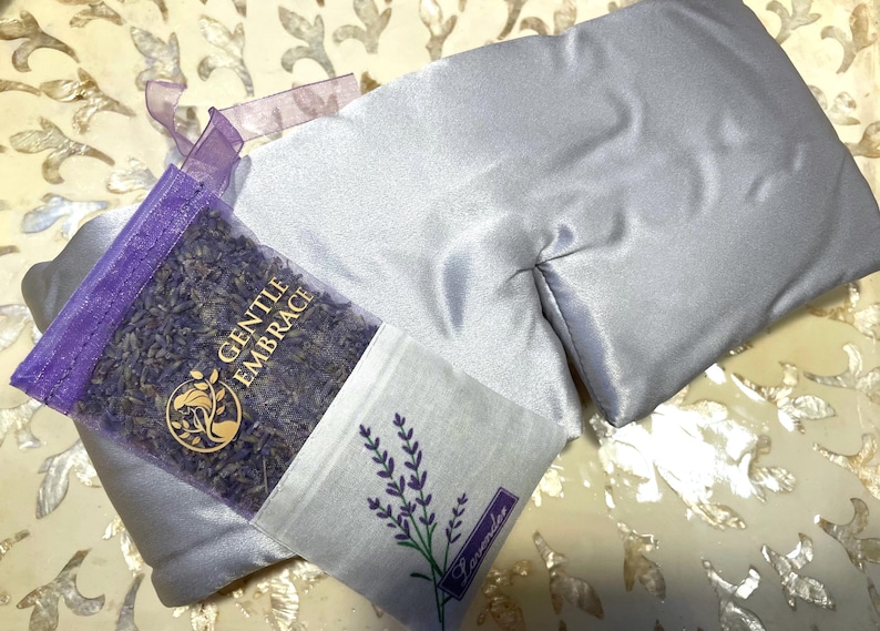 May include: A light grey satin eye pillow and a small sachet filled with dried lavender. The sachet has a lavender sprig illustration and the word "Lavender". A clear bag of lavender buds is visible with the text "GENTLE EMBRACE".