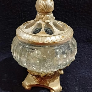 Pode incluir: Um frasco de vidro antigo com tampa e base decorativas prateadas. O frasco tem um corpo de vidro transparente e texturizado e uma tampa com um design floral. A base tem um design de três pés. O frasco é provavelmente usado para guardar pequenos objetos.