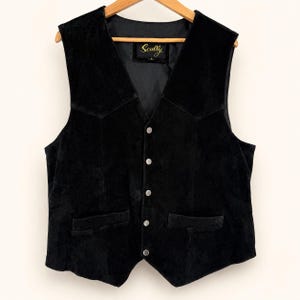 Può includere: Un gilet in camoscio nero con scollo a V, cinque bottoni in metallo argentato sul davanti e due tasche a filetto. L'etichetta interna riporta il nome del marchio Scully in caratteri dorati su uno sfondo nero.