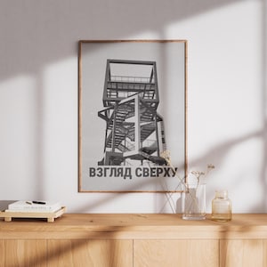 May include: Framed black and white architectural print featuring a multi-level structure with stairs and platforms. The print has the text "ВЗГЛЯД СВЕРХУ" at the bottom. The artwork is displayed on a light-colored wall above a wooden cabinet.