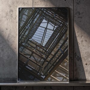 Może przedstawiać: Oprawiona fotografia struktury architektonicznej, widziana z dołu. Obraz przedstawia złożony układ metalowych belek i podpór, z widokiem na niebo przez centralny otwór. Dzieło sztuki jest eksponowane na tle betonowej ściany.