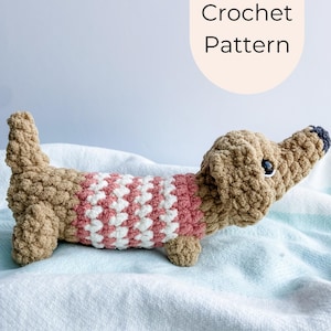 May include: A crocheted dachshund dog in tan and pink, wearing a pink and white striped sweater. The dog has black eyes and a black nose. The image includes the text "PDF Crochet Pattern" in a cream-colored oval.