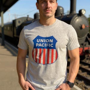 May include: A gray t-shirt featuring the Union Pacific logo in a shield design with blue, red, and white colors. The shirt is worn by a person standing in front of a vintage train. The person is wearing blue jeans and a black watch.