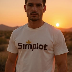 Può includere: Una maglietta bianca girocollo con la scritta "Simplot" in lettere marroni e un disegno a forma di foglia gialla sopra la parola. La maglietta è indossata da una persona davanti a un tramonto.