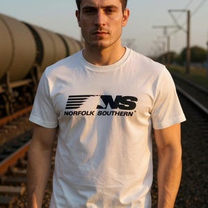 以下が含まれることがあります： 黒い「NS NORFOLK SOUTHERN」の文字が入った白いTシャツ。Tシャツはクルーネックで半袖です。背景には電車と線路があり、屋外で撮影された画像です。