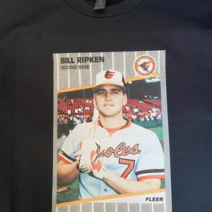 Pode incluir: Camiseta preta com estampa de cartão de beisebol de Bill Ripken, segunda base do Baltimore Orioles. O cartão mostra Ripken em seu uniforme do Orioles, segurando um bastão. A palavra "FLEER" está impressa na parte inferior.