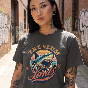 May include: Dark gray t-shirt featuring a shark graphic with the text "The Slum Lords" in a retro design. The shark is blue and white, with orange and yellow accents. The shirt is worn by a person with tattoos.