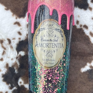 May include: A cylindrical tumbler with a clear lid, featuring a black base covered in multi-colored glitter. The top has a pink drip effect. A central label reads: THE APOTHECARY EST. 1766, Ensnare the Soul, AMORTENTIA, For Afflictions of the Heart.