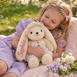 Può includere: Un peluche a forma di coniglio color crema con orecchie lunghe, che indossa un vestitino a stampa floreale e una cuffietta abbinata. Il nome Sophia è ricamato in corsivo rosa su una delle orecchie del coniglio. Il giocattolo ha piccoli occhi neri.