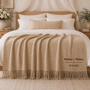 Könnte beinhalten: Eine beigefarbene Strickdecke mit Fransen, die mit dem gestickten Text Andrew + Monica, einem Herzsymbol und dem Datum 25.05.2027 versehen ist. Die Decke liegt auf einem Bett mit weißer Bettwäsche, einem passenden Lendenkissen und einem Holzkopfteil.