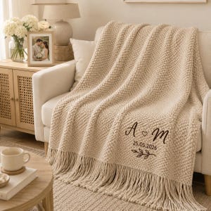 Può includere: Una coperta lavorata a maglia color beige con frange, caratterizzata da un ricamo marrone scuro con le lettere A e M, un piccolo simbolo a cuore, la data 25.05.2026 e un motivo decorativo a ramo con foglie nell'angolo in basso a destra.