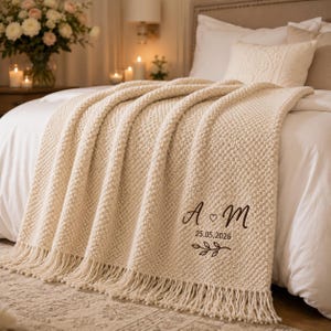 May include: A cream-coloured knitted throw blanket with a textured bobble pattern and fringe edges. The blanket features dark brown embroidered text that reads A heart M, the date 25.05.2026, and a small leafy branch graphic design.