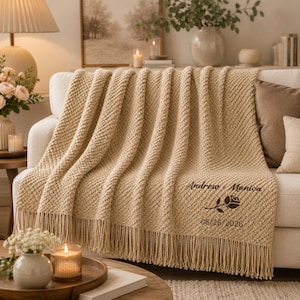 May include: A beige knit throw blanket with fringe trim, featuring the dark brown embroidered text Andrew + Monica, a rose graphic, and the date 08/26/2026. The blanket has a textured, raised stitch pattern and is draped over a white sofa.