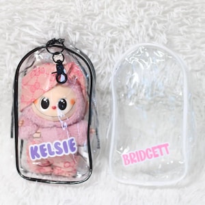 May include: A pink plush doll in a clear plastic case with the name "KELSIE" in purple letters. Beside it is an empty clear plastic case with the name "BRIDGETT" in pink letters. Both cases are on a white, fluffy surface.