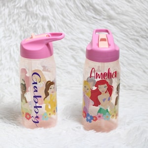 May include: Two clear plastic water bottles with pink lids and flip-up spouts. Each bottle features colorful cartoon princess graphics and floral accents at the base. The left bottle has the name Gabby printed vertically, and the right bottle has the name Amelia.