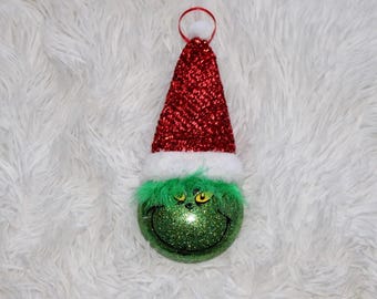 Puede incluir: Un adorno navideño con una cara verde brillante con una sonrisa traviesa, ojos amarillos y pelaje verde. El adorno está rematado con un gorro de Papá Noel rojo brillante con ribete blanco.