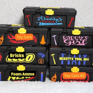 Pode incluir: Caixas de ferramentas pretas com fechos amarelos e vários decalques. Os decalques incluem frases como "Daddy's Little Helper", "Toy Cars" e "Beckett's Tool Box".