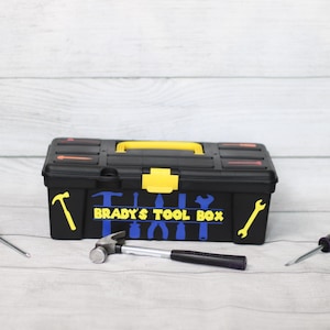 Puede incluir: Caja de herramientas negra con detalles amarillos y azules, con la inscripción "BRADY'S TOOL BOX". La caja de herramientas tiene un asa y un cierre amarillos. Dos destornilladores y un martillo están colocados cerca de la caja.