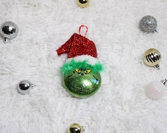 Puede incluir: Un adorno navideño verde brillante con forma de personaje, con un gorro de Papá Noel rojo y un ribete de piel verde. Varios otros adornos decorativos en plata y oro están esparcidos sobre una superficie blanca y esponjosa.