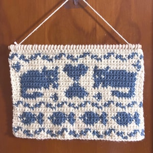 May include: A cream-coloured, rectangular crochet wall hanging with blue cat and heart motifs. It is suspended by a wooden dowel and white string. The design includes repeating patterns of fish and geometric shapes.