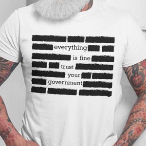 Può includere: T-shirt bianco con la scritta "everything is fine trust your government" in nero, in stile oscurato. La maglietta è indossata da una persona con tatuaggi e barba bianca.