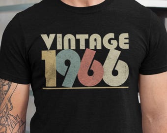 Peut inclure: T-shirt noir avec l'inscription "VINTAGE 1966" dans un style rétro. Le texte est composé de beige, jaune, bleu et rouge. Le t-shirt est à col rond et semble être en coton.