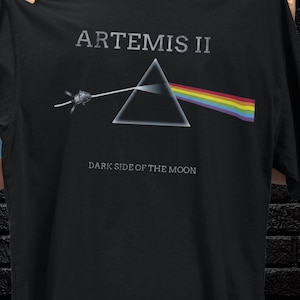 May include: Black t-shirt featuring the text "ARTEMIS II" above a triangle with a rainbow beam of light. Below the triangle is the text "DARK SIDE OF THE MOON". The design includes a spacecraft.