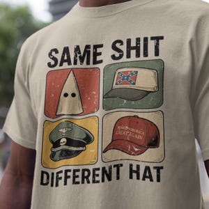Può includere: T-shirt beige con la scritta "SAME SHIT DIFFERENT HAT" sopra quattro immagini di cappelli. Le immagini includono un cappuccio del KKK, un cappello con la bandiera confederata, un cappello militare e un cappello "Make America Great Again".