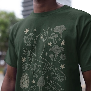 Può includere: T-shirt verde scuro con un design color crema di rane, funghi e stelle. La grafica include varie pose di rane e diversi tipi di funghi, creando un tema ispirato alla natura.