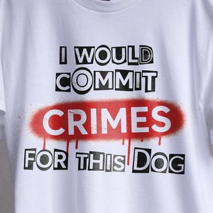 Può includere: T-shirt bianca con la scritta "I WOULD COMMIT CRIMES FOR THIS DOG" in nero, con "CRIMES" in rosso stile spray. La maglietta è appesa a un gancio.