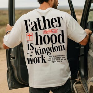 Può includere: T-shirt color crema con le parole "Father is hood kingdom work" in nero, con una croce rossa e stelle. Il testo "Proverbi 22:6" è sopra la parola "hood". La maglietta ha anche un versetto biblico stampato.