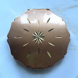 May include: A vintage, copper-toned compact with a scalloped edge. The compact features a starburst design in the center, with etched lines radiating outwards. The clasp is visible at the bottom. A classic beauty accessory.