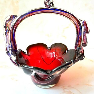 Può includere: Un cestino in vetro fatto a mano con interno rosso e bordo scuro. Il manico è trasparente con accenti blu e rossi. Il cestino ha una forma decorativa, a forma di fiore, e un piccolo elemento decorativo in vetro sulla parte superiore del manico.