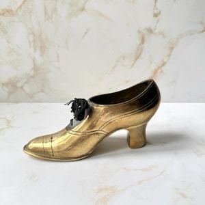 May include: A vintage brass shoe figurine with a high heel and black shoelaces. The shoe has a classic design with detailed stitching and a polished finish. The shoe is a decorative item.